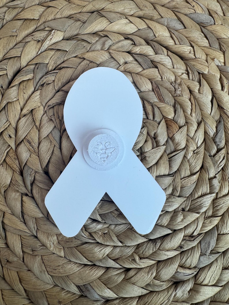 May include: A white ribbon-shaped pin with a circular emblem featuring a bee design. The pin is set against a woven, textured background, creating a contrast in textures and colors. The pin is likely a symbol of awareness or support.