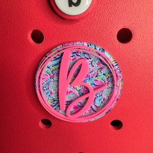 Könnte beinhalten: Ein rosa Buchstabe "B" mit weißem Hintergrund und einem bunten Paisley-Muster. Der Buchstabe ist von einem weißen Kreis mit rosa Rand umgeben.