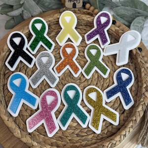 May include: A collection of awareness ribbon-shaped stickers in various colors, including black, green, yellow, purple, silver, orange, blue, pink, teal, and gold. Each ribbon has a glittery texture and a white border. The image also includes a small wooden sign that says "bee happy".