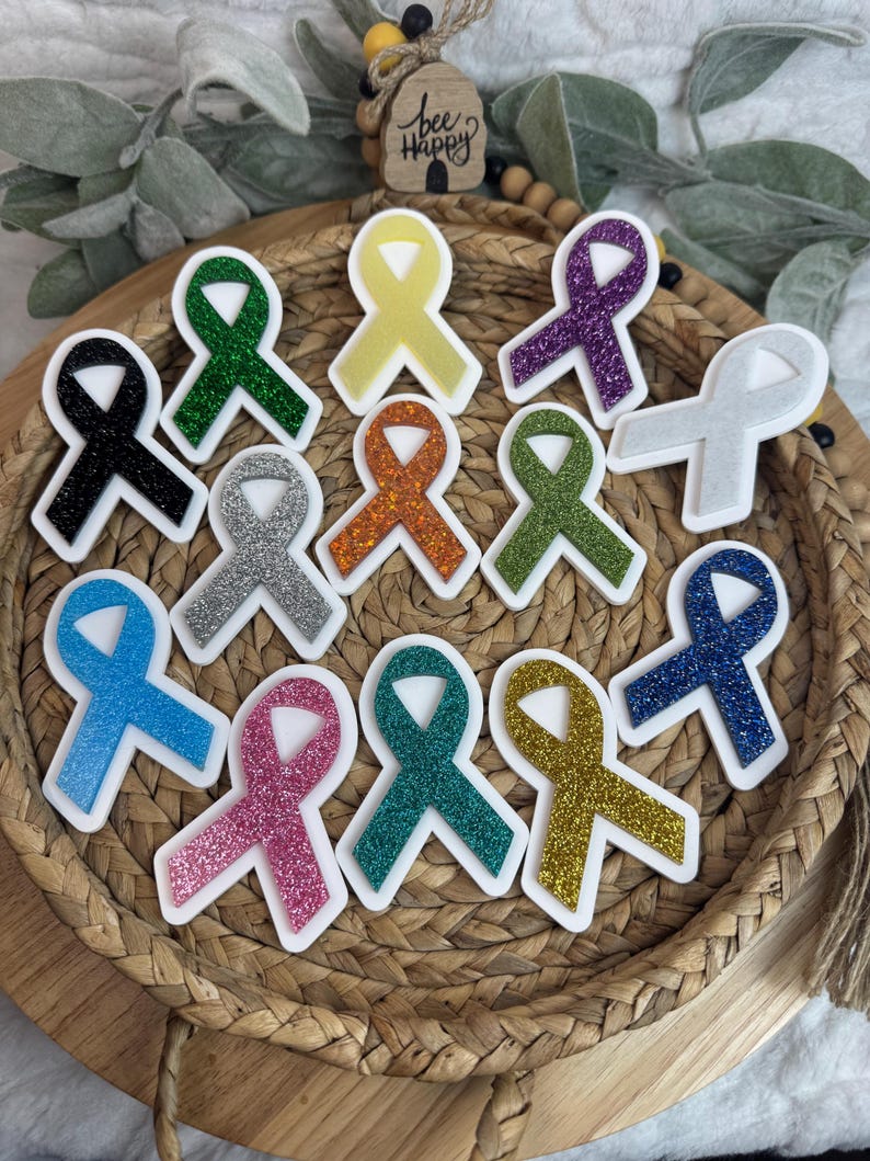 May include: A collection of awareness ribbon-shaped stickers in various colors, including black, green, yellow, purple, silver, orange, blue, pink, teal, and gold. Each ribbon has a glittery texture and a white border. The image also includes a small wooden sign that says "bee happy".