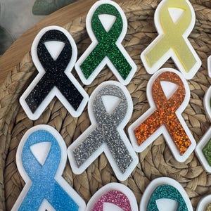 May include: A collection of colorful awareness ribbon-shaped acrylic charms with glitter accents. The ribbons are in various colors, including black, green, silver, orange, blue, pink, yellow, and teal, each with a white border. The charms are arranged on a woven surface.