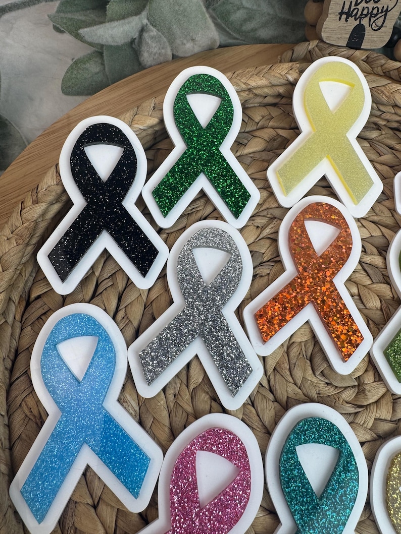 May include: A collection of colorful awareness ribbon-shaped acrylic charms with glitter accents. The ribbons are in various colors, including black, green, silver, orange, blue, pink, yellow, and teal, each with a white border. The charms are arranged on a woven surface.