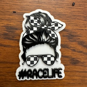 Op de afbeelding: Een zwart-wit geruit ontwerp op een vrouwenhaarbol, een zonnebril en een bandana. De tekst #RACELIFE staat onder de afbeelding.
