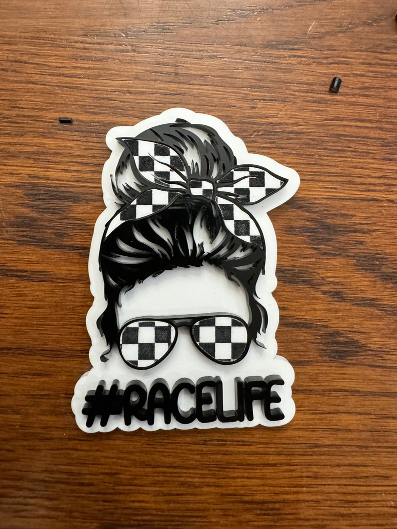 Op de afbeelding: Een zwart-wit geruit ontwerp op een vrouwenhaarbol, een zonnebril en een bandana. De tekst #RACELIFE staat onder de afbeelding.