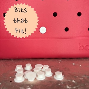 May include: A close-up of white plastic buttons, also known as "jibbitz", that can be used to customize a pair of shoes. The buttons are scattered on a table. The text "Bits that Fit!" is visible in the background.