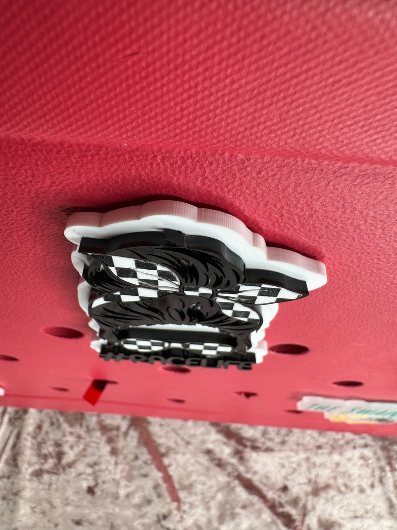 Op de afbeelding: Witte en zwarte 3D-geprinte bedel met een geruit patroon en de tekst "#racelife" op een rode achtergrond.