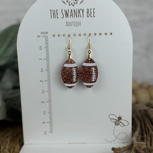 May include: A pair of gold tone earrings with brown glitter football charms. The earrings are displayed on a white card with the text "The Swanky Bee Boutique".