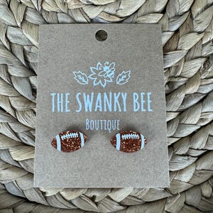 May include: A pair of brown glitter football earrings with white stitching. The earrings are on a card that says "The Swanky Bee Boutique".