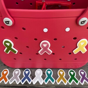 May include: A bright pink tote bag with several ribbon-shaped charms attached. The charms are in various colors, including green, pink, and yellow. Additional ribbon charms in multiple colors are lined up below the bag.