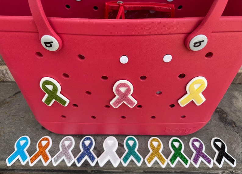May include: A bright pink tote bag with several ribbon-shaped charms attached. The charms are in various colors, including green, pink, and yellow. Additional ribbon charms in multiple colors are lined up below the bag.