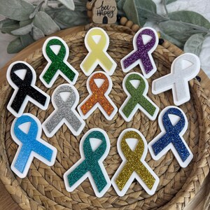 May include: A collection of colorful awareness ribbon stickers in various colors, including black, green, yellow, purple, white, orange, and blue. Each ribbon has a glittery texture and a white border. The ribbons are arranged in a woven basket.