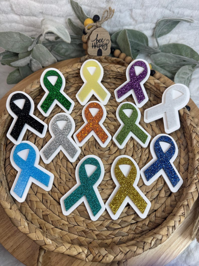 May include: A collection of colorful awareness ribbon stickers in various colors, including black, green, yellow, purple, white, orange, and blue. Each ribbon has a glittery texture and a white border. The ribbons are arranged in a woven basket.