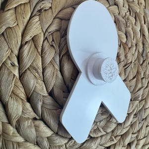 May include: A white awareness ribbon with a circular base, possibly a pin or decoration. The ribbon is made of a solid, opaque material and is set against a woven, textured background.
