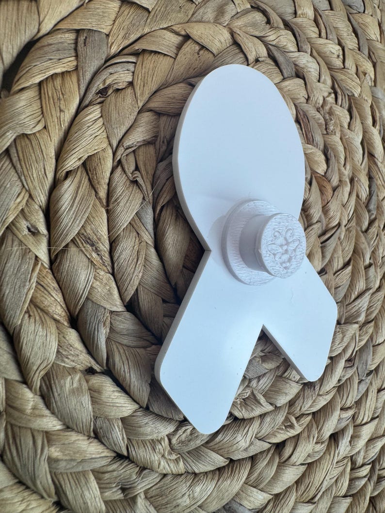 May include: A white awareness ribbon with a circular base, possibly a pin or decoration. The ribbon is made of a solid, opaque material and is set against a woven, textured background.