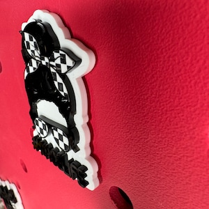 Op de afbeelding: Een witte en zwarte 3D-geprinte sticker met een geruit bandana, een zonnebril en de tekst "#badlife" op een rode achtergrond.