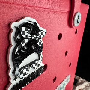 Op de afbeelding: Een rode draagtas met een wit-zwarte geruite sticker van een persoon met een zonnebril en een hoofdband. De sticker zegt "#racelife".