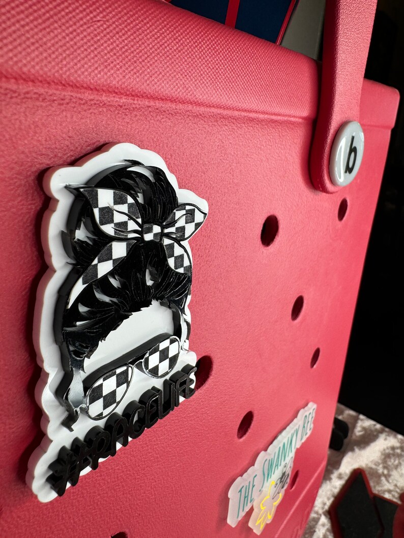 Op de afbeelding: Een rode draagtas met een wit-zwarte geruite sticker van een persoon met een zonnebril en een hoofdband. De sticker zegt "#racelife".