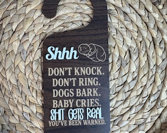 May include: Dark brown wooden door hanger with a curved top. The text "Shhh" and a baby illustration are at the top. Below, white text reads: "DON'T KNOCK. DON'T RING. DOGS BARK. BABY CRIES. SHIT GETS REAL. YOU'VE BEEN WARNED."