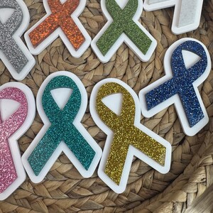 May include: Assortment of glittery awareness ribbon-shaped stickers in various colors, including pink, orange, green, teal, gold, and blue. Each ribbon has a white border. The stickers are arranged on a woven basket.