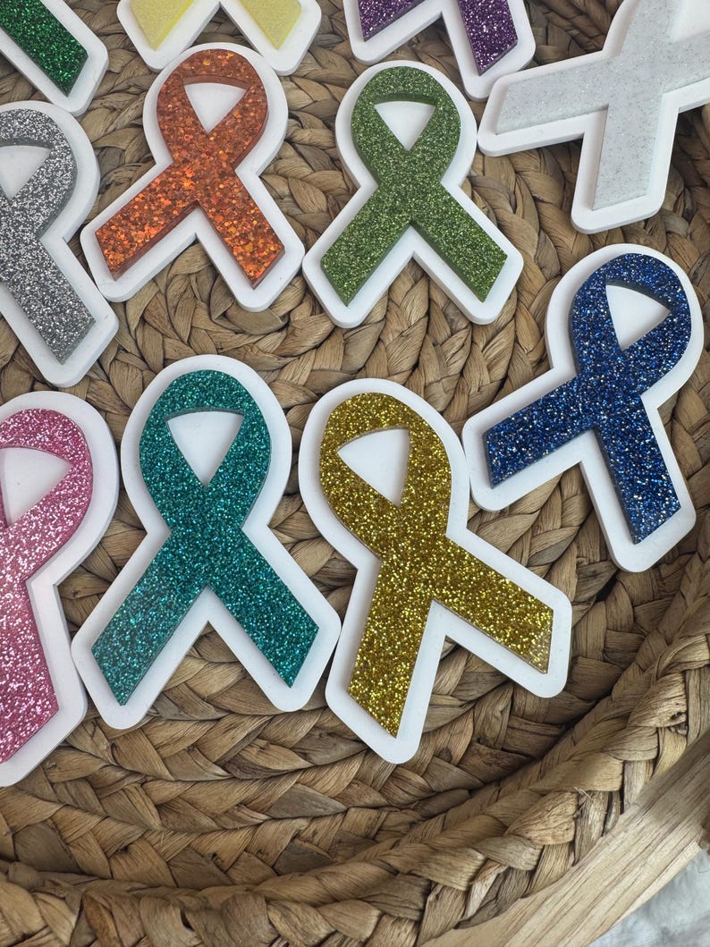 May include: Assortment of glittery awareness ribbon-shaped stickers in various colors, including pink, orange, green, teal, gold, and blue. Each ribbon has a white border. The stickers are arranged on a woven basket.