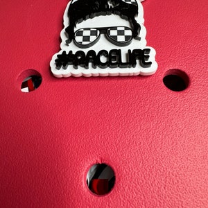 Op de afbeelding: Witte sticker met een zwart-wit geruit patroon, een zwart silhouet van een persoon met een zonnebril en een bandana, en de tekst "#RACELIFE".