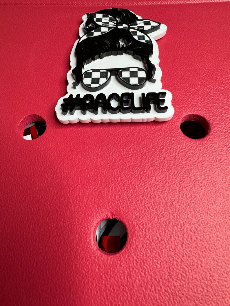 Op de afbeelding: Witte sticker met een zwart-wit geruit patroon, een zwart silhouet van een persoon met een zonnebril en een bandana, en de tekst "#RACELIFE".