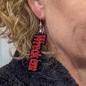May include: Red glitter earrings with the word "Wrecked" written on them. The earrings are hanging from a person's ear.