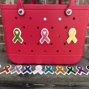 May include: A bright pink tote bag with a variety of colorful awareness ribbon charms. The bag has a series of holes and white accents. Below the bag are several ribbon charms in various colors, including blue, orange, and purple.