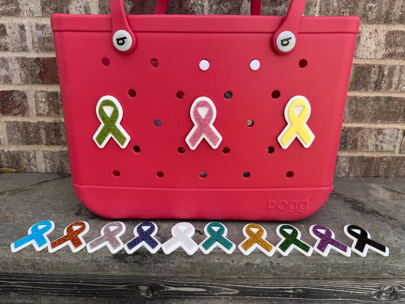 May include: A bright pink tote bag with a variety of colorful awareness ribbon charms. The bag has a series of holes and white accents. Below the bag are several ribbon charms in various colors, including blue, orange, and purple.