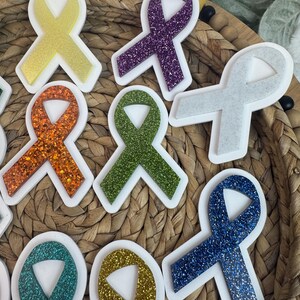 May include: A collection of glittery awareness ribbon stickers in various colors, including yellow, purple, orange, green, gray, blue, and teal. Each ribbon has a white border. The stickers are arranged on a woven surface.