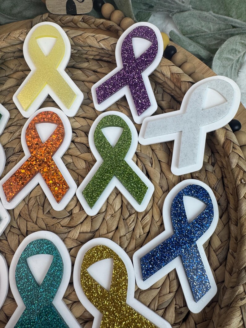 May include: A collection of glittery awareness ribbon stickers in various colors, including yellow, purple, orange, green, gray, blue, and teal. Each ribbon has a white border. The stickers are arranged on a woven surface.