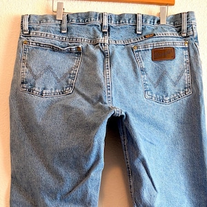 Puede incluir: Vaqueros de mezclilla azul claro con un dise&ntilde;o cl&aacute;sico de cinco bolsillos. Los vaqueros muestran el logotipo de Wrangler en los bolsillos traseros y un parche de cuero marr&oacute;n. Los vaqueros cuelgan de una percha de madera.