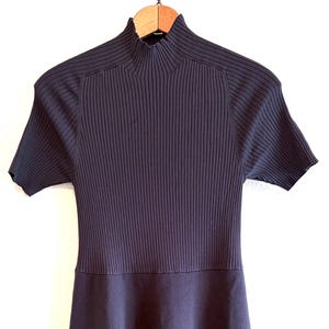Può includere: Abito blu navy a maniche corte con corpetto a coste e gonna svasata. L'abito ha un collo alto ed è appeso a una gruccia di legno su sfondo bianco. L'abito è realizzato in morbido tessuto a maglia.