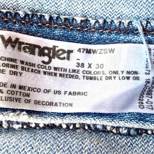 Puede incluir: Primer plano de una etiqueta de vaqueros Wrangler. La etiqueta blanca tiene texto negro, mostrando la marca, la talla 38 x 30 y las instrucciones de lavado. La tela es 100% algod&oacute;n, fabricada en M&eacute;xico con tejido estadounidense.