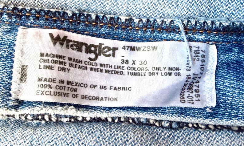 Puede incluir: Primer plano de una etiqueta de vaqueros Wrangler. La etiqueta blanca tiene texto negro, mostrando la marca, la talla 38 x 30 y las instrucciones de lavado. La tela es 100% algod&oacute;n, fabricada en M&eacute;xico con tejido estadounidense.