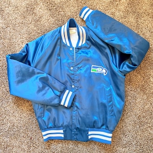 Puede incluir: Una chaqueta universitaria de satén azul brillante con cuello, puños y dobladillo acanalados a rayas blancas y azules. La chaqueta tiene un cierre frontal con botones a presión y un parche con el logotipo de los Seattle Seahawks en el pecho.