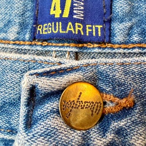 Puede incluir: Primer plano de unos vaqueros azules con un bot&oacute;n de lat&oacute;n y una etiqueta azul. La etiqueta dice "47 MWZ REGULAR FIT" en letras amarillas. El bot&oacute;n es redondo con un dise&ntilde;o decorativo.