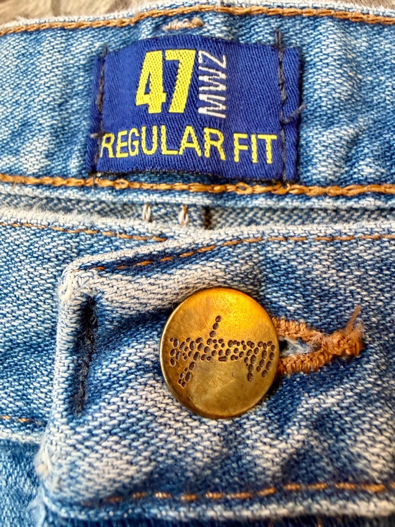Puede incluir: Primer plano de unos vaqueros azules con un bot&oacute;n de lat&oacute;n y una etiqueta azul. La etiqueta dice "47 MWZ REGULAR FIT" en letras amarillas. El bot&oacute;n es redondo con un dise&ntilde;o decorativo.