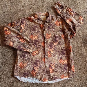 Peut inclure: Chemise boutonnée à manches longues avec un motif camouflage représentant des feuilles et des cerfs bruns, orange et verts. La chemise a un col boutonné et une poche poitrine. Le tissu semble être un mélange de coton.