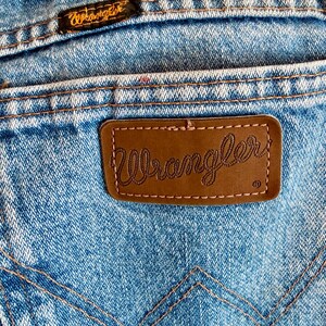 Puede incluir: Primer plano del bolsillo trasero de unos vaqueros azules. El parche de cuero marr&oacute;n presenta el nombre de la marca "Wrangler" en cursiva. Los vaqueros tienen costuras naranjas y una peque&ntilde;a etiqueta "Wrangler" cerca de la cintura.