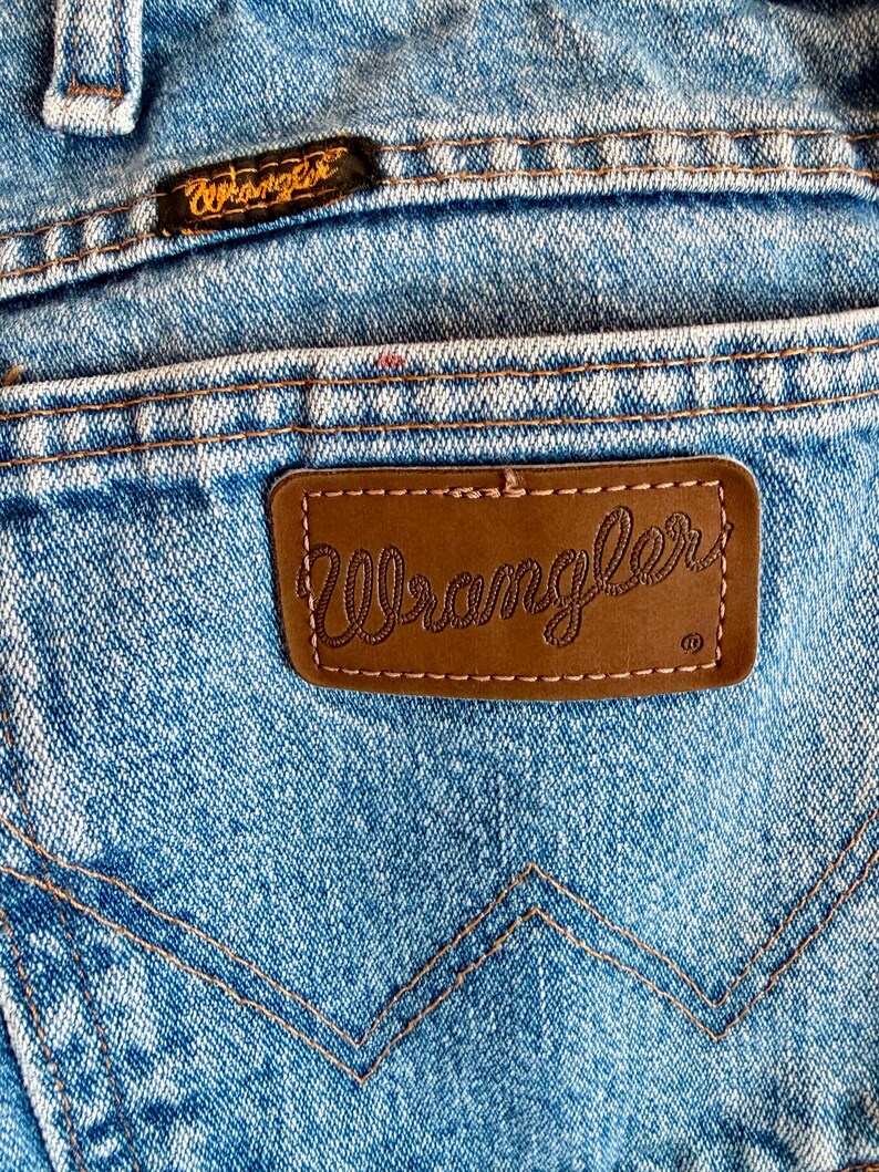 Puede incluir: Primer plano del bolsillo trasero de unos vaqueros azules. El parche de cuero marr&oacute;n presenta el nombre de la marca "Wrangler" en cursiva. Los vaqueros tienen costuras naranjas y una peque&ntilde;a etiqueta "Wrangler" cerca de la cintura.