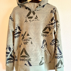 Op de afbeelding: Een lichtgroene Reebok hoodie met een herhaald patroon van het Reebok logo en tekst in zwart. De hoodie heeft een voorzak, een capuchon met een donkergroene voering en geribde manchetten en zoom.