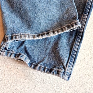 Puede incluir: Primer plano de unos vaqueros de mezclilla azul claro. La imagen destaca el dobladillo doblado y las costuras detalladas a lo largo de las costuras. La mezclilla tiene una apariencia descolorida, lo que sugiere desgaste. Los vaqueros est&aacute;n sobre una superficie texturizada blanquecina.