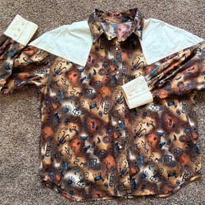 May include: A long-sleeve button-down shirt featuring a brown and orange gradient background with a repeating pattern of blue and black stylized figures, animals, and geometric shapes. The shirt has cream-colored shoulder yokes and cuffs.