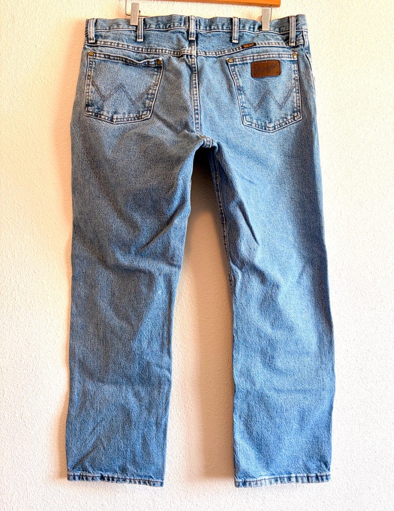 Puede incluir: Vaqueros de mezclilla azul claro con un dise&ntilde;o cl&aacute;sico de cinco bolsillos. Los vaqueros muestran el logotipo de Wrangler en los bolsillos traseros y un parche de cuero marr&oacute;n. Los vaqueros cuelgan de una percha de madera.