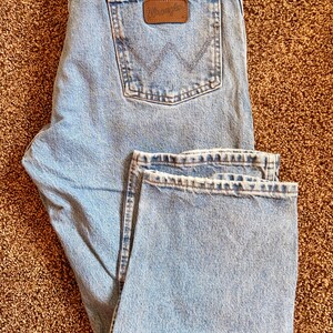 Puede incluir: Vaqueros de mezclilla azul claro con el logo de Wrangler en el bolsillo trasero. Los vaqueros est&aacute;n doblados, mostrando la parte delantera y trasera. Los vaqueros tienen un dise&ntilde;o cl&aacute;sico de cinco bolsillos y corte recto.