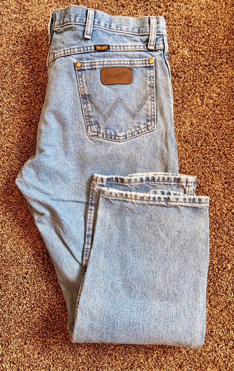 Puede incluir: Vaqueros de mezclilla azul claro con el logo de Wrangler en el bolsillo trasero. Los vaqueros est&aacute;n doblados, mostrando la parte delantera y trasera. Los vaqueros tienen un dise&ntilde;o cl&aacute;sico de cinco bolsillos y corte recto.