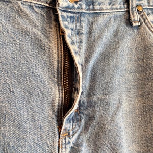 Puede incluir: Primer plano de unos vaqueros de mezclilla azul claro. Los vaqueros tienen una cremallera visible, un cierre de bot&oacute;n y remaches de color lat&oacute;n. La tela muestra una textura desgastada, con costuras visibles y la marca "AT" en la etiqueta.