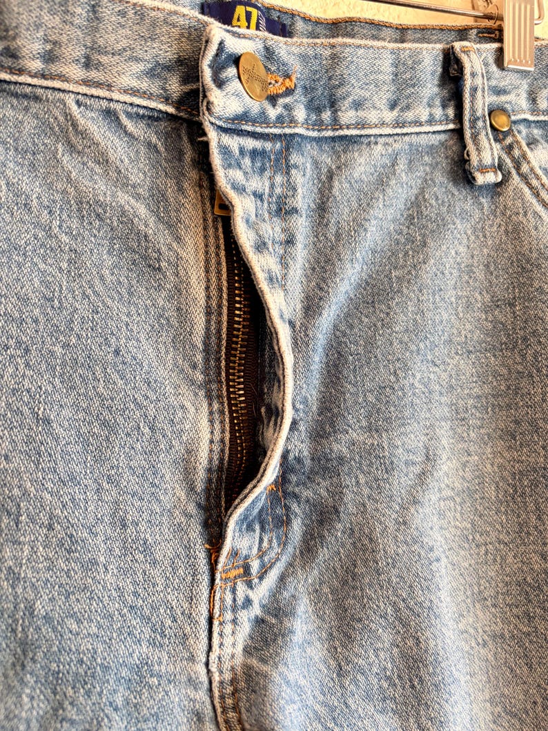 Puede incluir: Primer plano de unos vaqueros de mezclilla azul claro. Los vaqueros tienen una cremallera visible, un cierre de bot&oacute;n y remaches de color lat&oacute;n. La tela muestra una textura desgastada, con costuras visibles y la marca "AT" en la etiqueta.