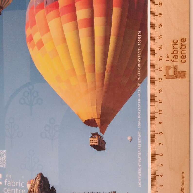 May include: A hot air balloon with orange and yellow stripes against a blue sky. The balloon is flying over a rocky landscape. A wooden ruler with the text "the fabric centre" is visible on the right side of the image. The ruler is marked in millimeters and inches.